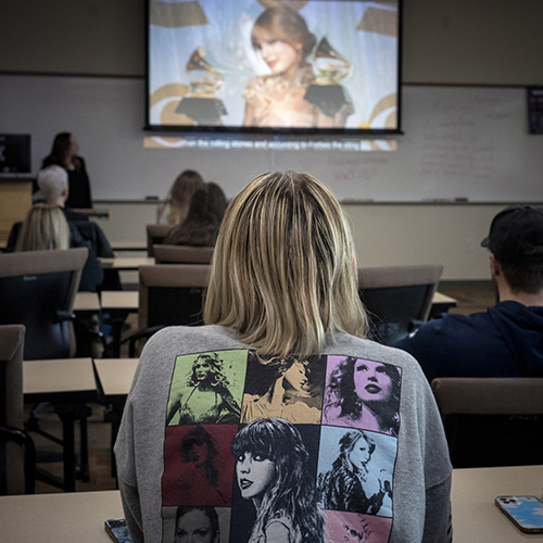 A blonde girl with a gray Taylor Swift Eras Tour sweatshirt sits in a classroom while attending a Taylor Swift Studies class.
