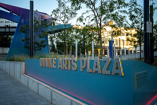 The image shows the Dumke Arts Plaza sign. The letters are white with a yellow outline and they are on a light blue background.
