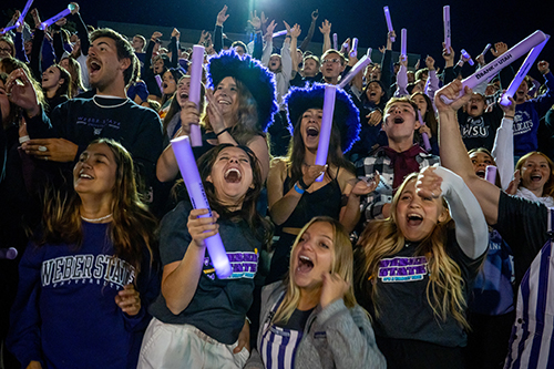 Weber State students cheer at the homecoming football game. They are all wearing their Weber State gear and holding purple light-up foam sticks.