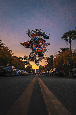 A dancer from Indigenous Soundscapes in Motion is jumping in the middle of an empty street while wearing his Native American cultural attire. The sun is setting in the background, and the sky has shades of blue, pink and yellow.