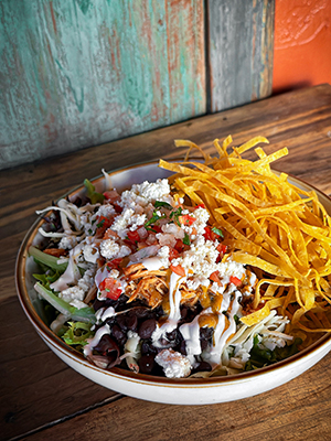 A plate of food from Sonora Grill is on a wooden table. The plate has lettuce, shredded chicken, black beans, pico de gallo, sour cream and corn tortilla strips. 