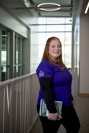 Nursing student Mandy Pellew wears purple scrubs and holds a nursing textbook 