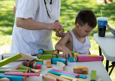 A child standing in front of an adult playing with blocks and wooden cars on a table. 